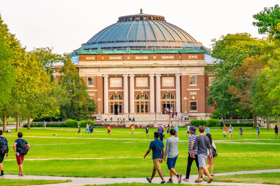 College students walking on green lawn with neoclassical red brick domed building in background on University of Illinois campus.